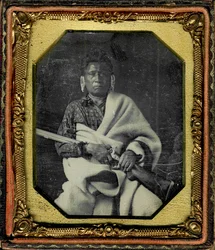 Porträt von Nacheninga [No Heart of Fear], Iowa. Nacheninga hält eine Waffe in den Händen und ist in eine dicke Decke gehüllt.